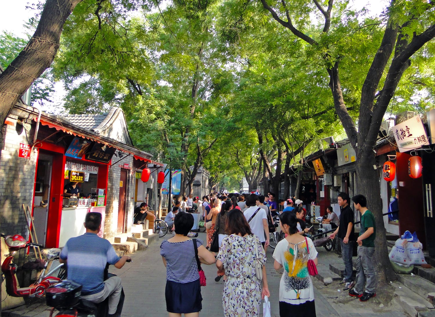 The Nanluoguxiang hutong in Beijing — a leafy old lane lined with small shops and cafés in brick buildings, red lanterns strung overhead