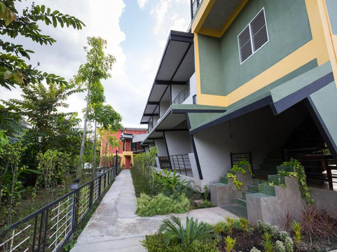 อาคารที่พักสองชั้นสีเขียว-เทาตัดเสาเหลือง ทางเดินคอนกรีตผ่านสวนที่ปลูกต้นไม้รอบตัว