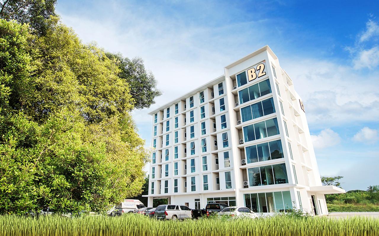 ตัวอาคารสีขาว 7 ชั้นของ B2 Phrae กลางวัน มีป้าย B2 และทุ่งนาด้านหน้า