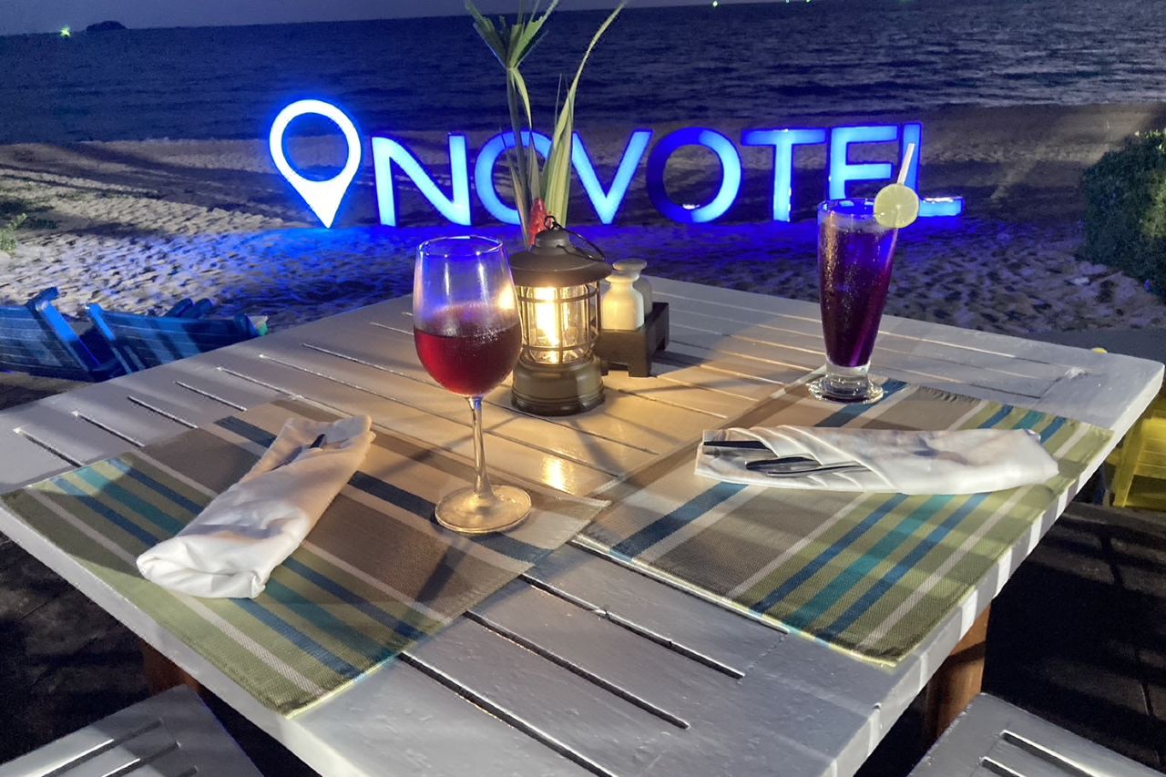โต๊ะอาหารริมหาดยามค่ำ กับป้าย Novotel เรืองแสงบนชายหาด